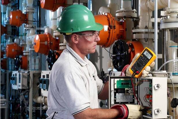 Tekniker måler spænding med et gult Fluke 87V multimeter i et industrielt anlæg.