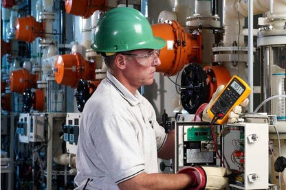 Tekniker måler spænding med et gult Fluke 87V multimeter i et industrielt anlæg.
